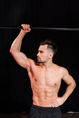 shirtless and muscular sportsman looking away and posing with hand on hip near horizontal bar isolated on black.