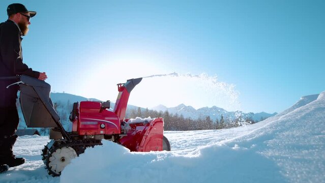 SLOW MOTION, COPY SPACE, LENS FLARE, CLOSE UP: Bearded man operates a snow blower on a sunny winter day. Male ski resort maintenance worker uses a snowblower on a picturesque sunny December day.