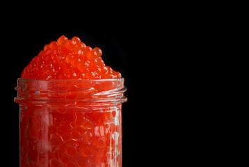 Open transparent glass jar with red salmon caviar on a black background. Free space for an inscription