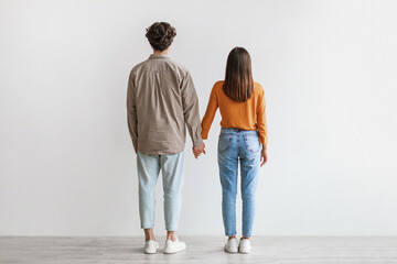 Back view of young Asian couple in casual wear standing, holding hands against white studio wall