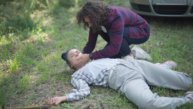 Unconscious knocked down woman lying on ground with man raising head checking pulse. Desperate frustrated Caucasian male driver and dead woman with car at background outdoors. Road accident