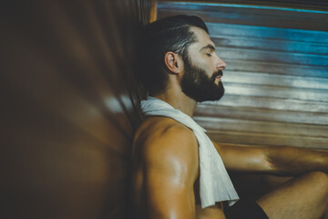 Beautiful healthy boy relaxing in a wooden sauna. Young bearded man resting after workout in the gym. Fitness male with tattoo and white towel, laying on the bench in the spa. Wellness, health concept - obrazy, fototapety, plakaty