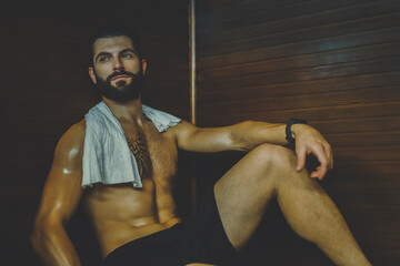 Fototapeta na wymiar Beautiful healthy boy relaxing in a wooden sauna. Young bearded man resting after workout in the gym. Fitness male with tattoo and white towel, laying on the bench in the spa. Wellness, health concept