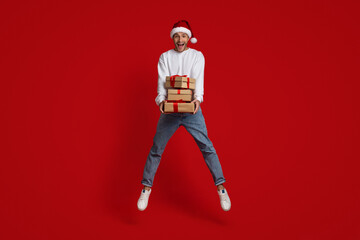 Fototapeta na wymiar Xmas Offer. Overjoyed Guy Wearing Santa Hat Jumping With Gifts In Hands