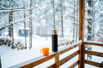 black matte thermos and orange mug with hot drink on window sill of the outdoor veranda. Hiking and picnic staff. Drinking warm tea or coffee in the winter frost.