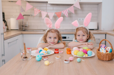 two little beautiful twin girls are sitting at a festive table while preparing for the Easter holiday watching the camera. close-up