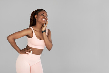 Young black woman in sports outfit wearing fitness tracker, posing and looking aside at empty space on grey background