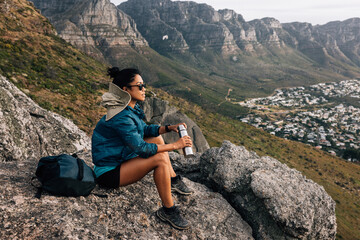Fototapeta premium Side view of woman sitting on a rock holding thermos. Female hiker relaxing while sitting on top of a mountain.