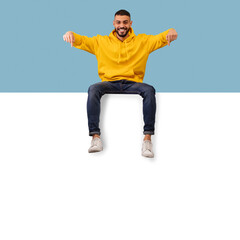 Cheerful arab man sitting on horizontal blank advertising board and pointing down at copy space, mockup
