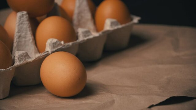 A chicken egg is rolling near the container. Chicken brown fresh raw eggs in a paper box. An egg carton with ten eggs. Lots of fresh raw chicken eggs. Omelette. 4k