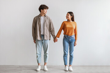 Full length of millennial Asian couple smiling and looking at each other, holding hands, standing against white wall - Powered by Adobe