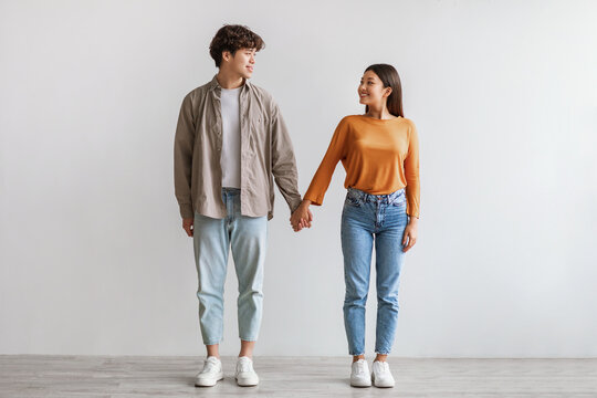 Full length of millennial Asian couple smiling and looking at each other, holding hands, standing against white wall