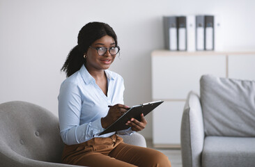 Happy black female psychologist taking notes in clipboard during consultation, smiling at camera in...