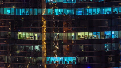 Office windows of a glazed skyscraper glow at night with city lights reflection aerial timelapse.