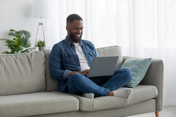 Smiling millennial black bearded male work on pc on sofa in living room interior, checking email in morning