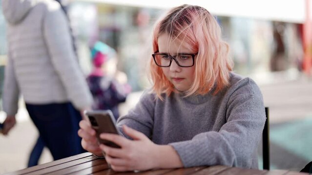 A teenage girl with glasses is sitting at a table in a street cafe and playing video games on her phone. Modern youth with a bright image in the style of hair color