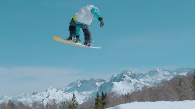 SLOW MOTION, CLOSE UP: Action shot of an athletic male tourist doing a 360 grab while exploring the ski resort fun park. Extreme snowboarder soars through the air and does a spinning grab trick.