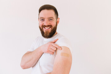 Portrait of smiling bearded man after getting a vaccine. Male holding up his white shirt sleeve and showing his arm with bandage after receiving vaccination. Concept of recommended inoculation	 - obrazy, fototapety, plakaty