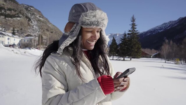 Latina woman using phone in winter forest with snowy landscape. Cheerful girl in the cold mountains types her cell phone.