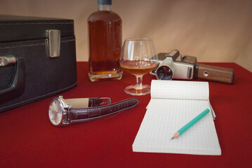 Bottle, glass, wrist watch, notebook and old movie camera on red table. Selective focus. Retro composition