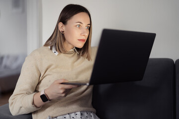 Caucasian Woman using laptop, staring at the screen surprised while sitting on the sofa at home in the evening living room - obrazy, fototapety, plakaty
