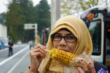 Portrait of smiling muslim woman eating roasted sweet corn at Hakonemachi-ko port station by Lake Ashi in Hakone, Japan with bus, street and tree in bokeh background. Smiling and happy expression.
