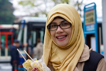 Close up portrait of muslim woman holding roasted sweet corn at Hakonemachi-ko port station by Lake Ashi in Hakone, Japan with bus and tree in bokeh background. Smiling and happy expression.