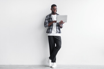Millennial black guy in casual wear using laptop computer for online work, education or communication against white wall