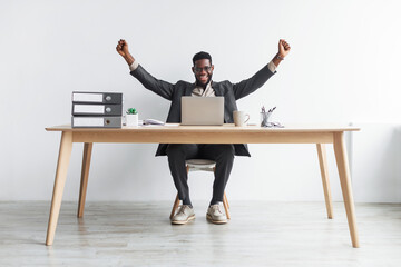 Full length of excited young black businessman sitting at desk with laptop, making YES gesture...