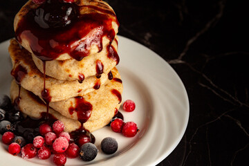 Several pancakes with berries in a stack on a white plate covered with sweet syrup