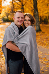 Young couple is walking in the sunny autumn park. Happy boy and girl are rolling themselves in the rug near yellow tree. Fallen yellow leaves, brith warm autumn weather. Autumn love story in park
