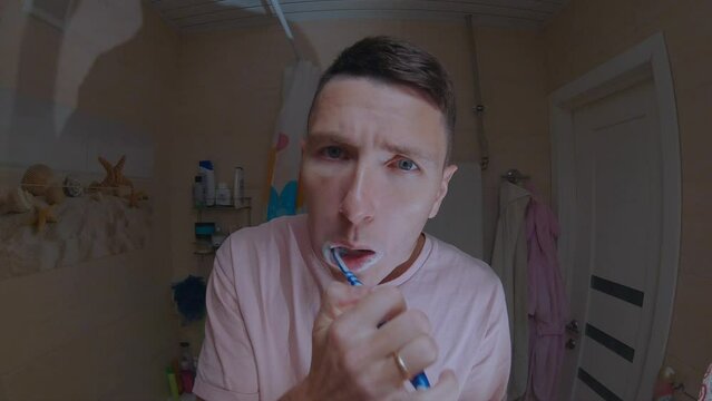A young man in a pink T-shirt brushes his teeth in front of a mirror.