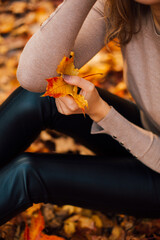 Beautiful girl is sitting on yellow leaves in the park. Attractive woman wearing beige sweater and black pants is walking in autumn park. Bright, warm sunny autumn day with yellow background.
