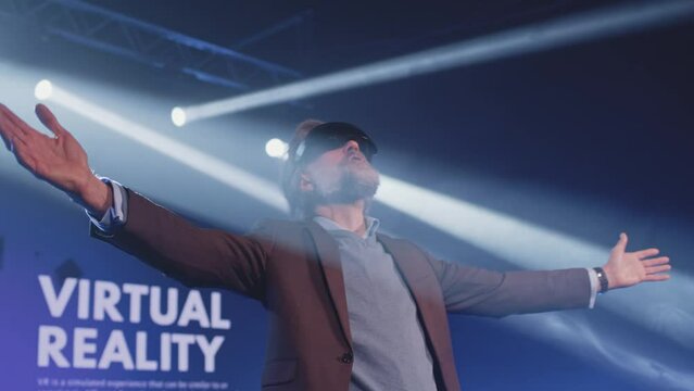 Happy male speaker in suit gesturing and demonstrating augmented reality glasses, to the audience at a Metaverse event on stage in front of an LED screen 3D objects in illuminated room