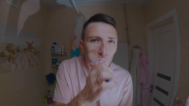 A young man in a pink T-shirt brushes his teeth in front of a mirror.