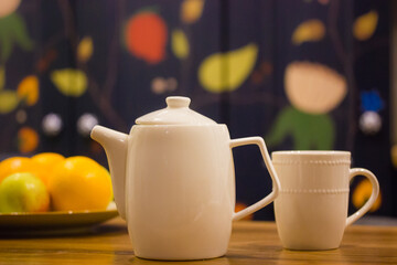 White ceramic teapot and a cup standing on a wooden table in the modern stylish kitchen against a colorful wall. Bowl of fruits. A fruity drink. Teapot, kettle for hot beverages. Kitchenware, utensil.