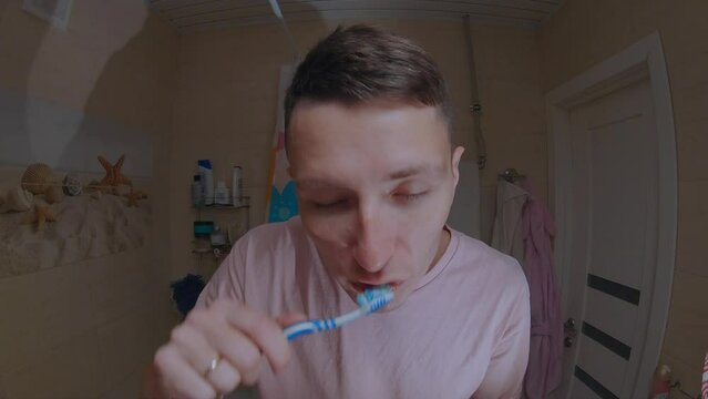 A young man in a pink T-shirt brushes his teeth in front of a mirror.