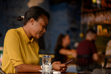 African american woman using a smartphone and drinking coffee while sitting in a cafe