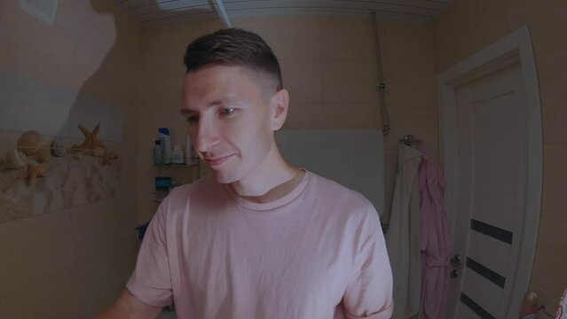 A young man in a pink T-shirt is applying toothpaste on a toothbrush in the bathroom