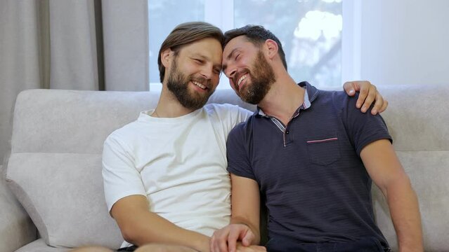 Gay couple smile and hug, spend time together relaxing while sitting on the sofa in the living room.