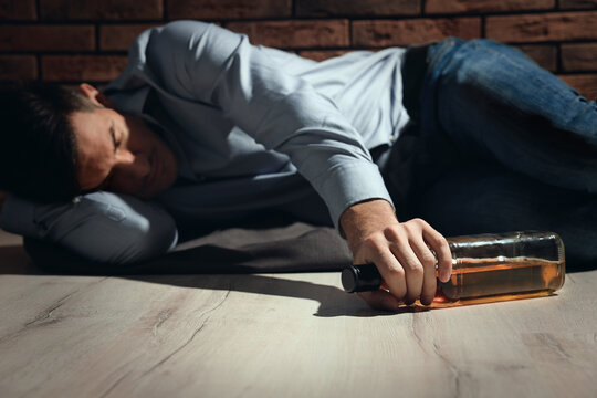 Addicted man with glass bottle of alcoholic drink lying on floor near red brick wall