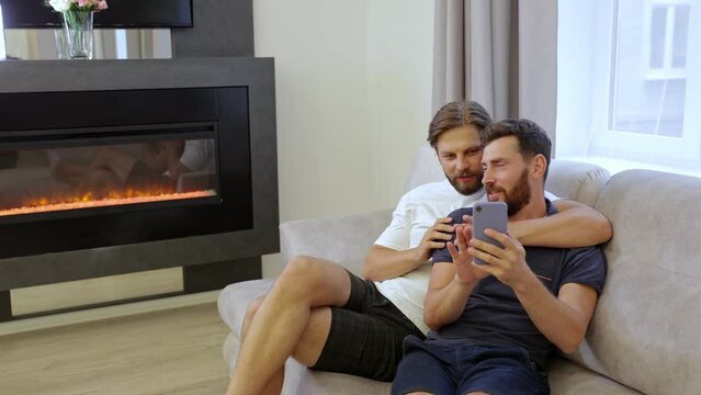 Cute male gay couple spending time at home. They lie on the couch and use a smartphone. They talk and shop online together.