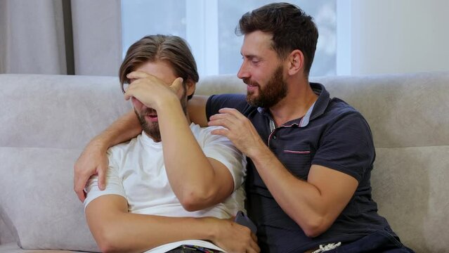 A man sits on the couch sad, crying, when a positive gay guy hugs and comforts his lover.
