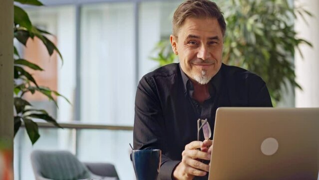 Business portrait - businessman sitting in in office working with laptop computer. Mature age, middle age, mid adult man in 50s with happy confident smile. Copy space.