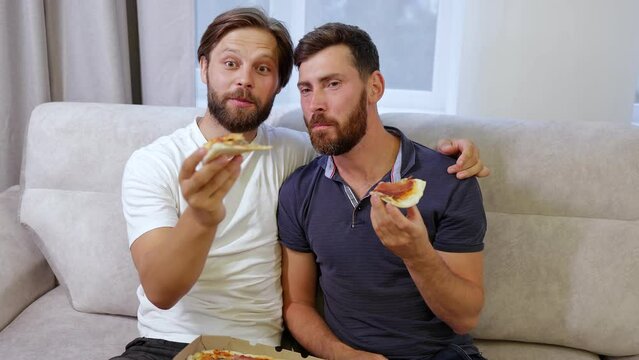 Loving gays eat slices of pizza. Two happy men sit at home on the sofa, eat pizza and enjoy. The right to same-sex love and the concept of free relationships