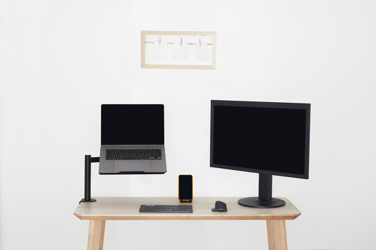 Wooden desk with a monitor, a laptop on a stand, a cell phone, a keyboard and a mouse. Black screens, white walls and a wooden frame with 3 photos.