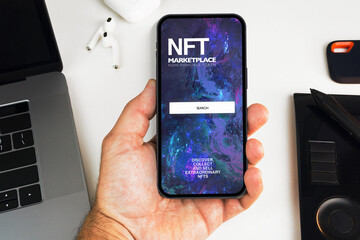 Man holding a smartphone with NFT (Non-Fungible Token) Marketplace on the screen on white background table. Office environment. Crypto art
