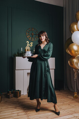 A beautiful brunette girl holds a delicious cake with the numbers 25 on her birthday against the backdrop of an interior with balloons. Festive photo.