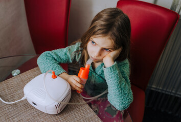 A girl, a child sits on a chair and makes inhalation with a special inhaler with a wire, inhales and exhales steam.