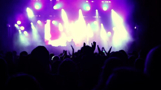 Illuminated by stage lights. Raised hands are visible. Concert crowd silhouettes in front of bright stage. People with raised hands. Concert crowd silhouettes in front of bright platform lights